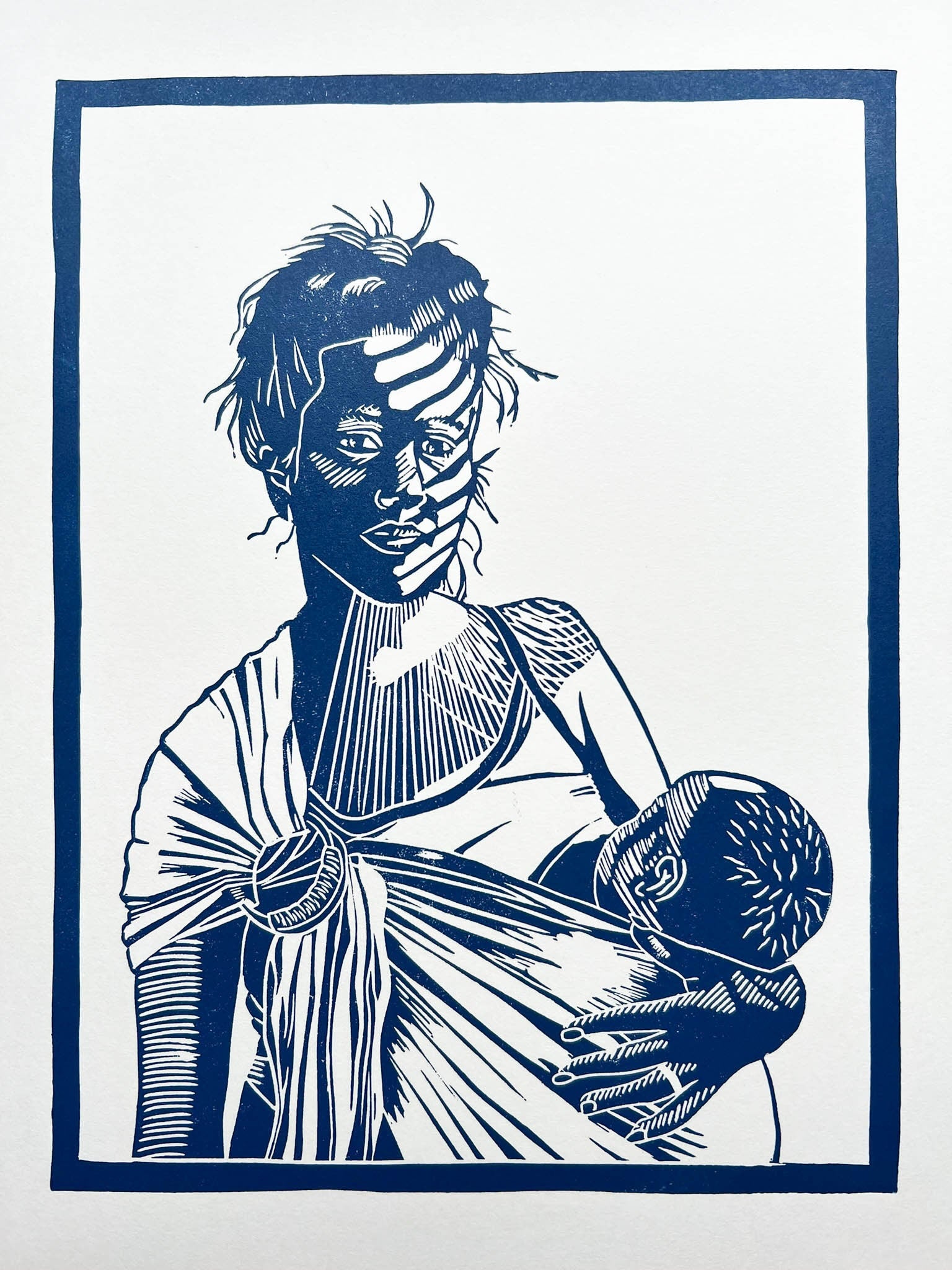 A blue lino print of a mother breastfeeding her baby in a ring sling with sunlight across her face through the blinds.