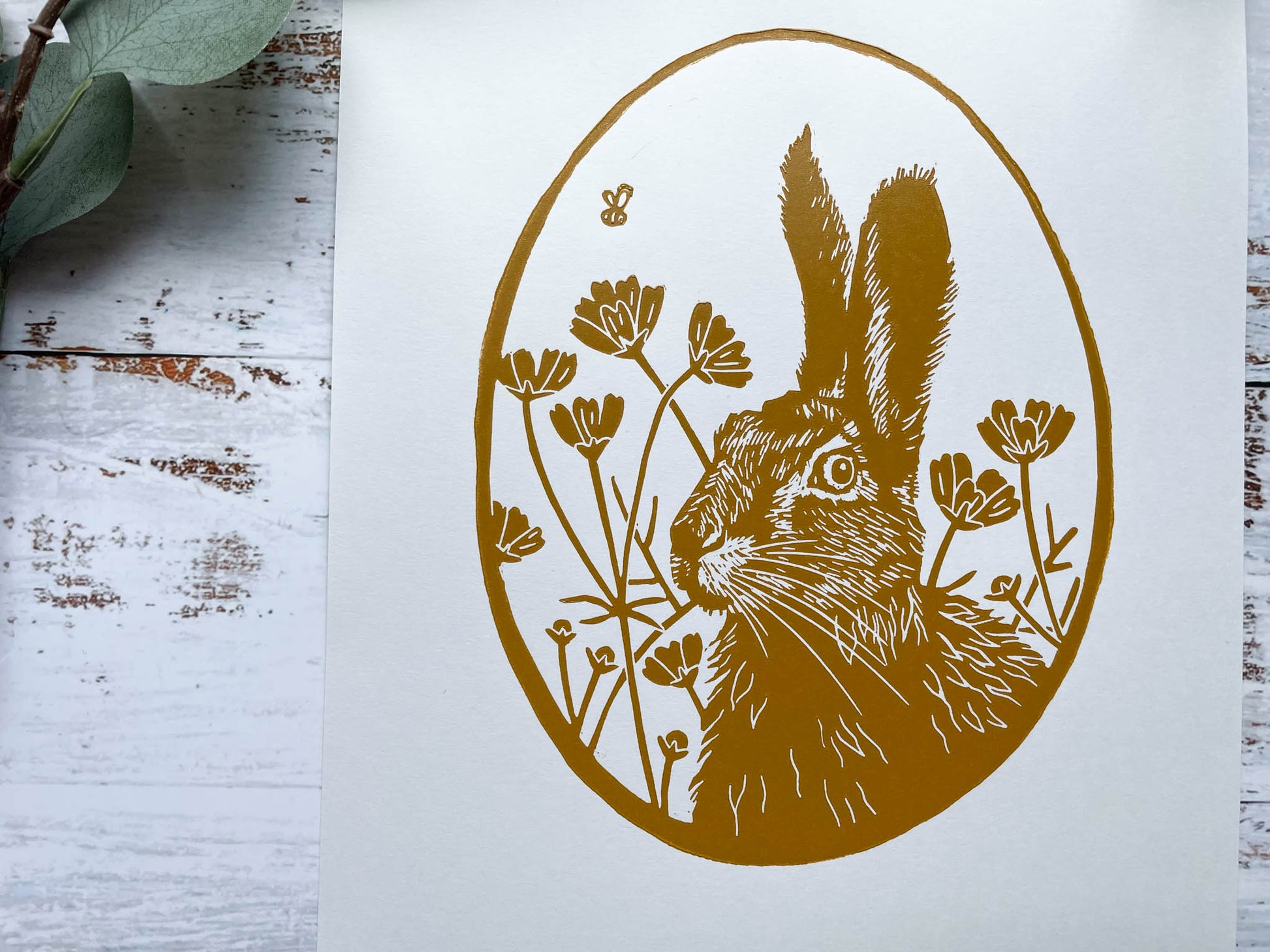 An oval lino print in yellow ochre of a hare surrounded by flowers on A4 paper